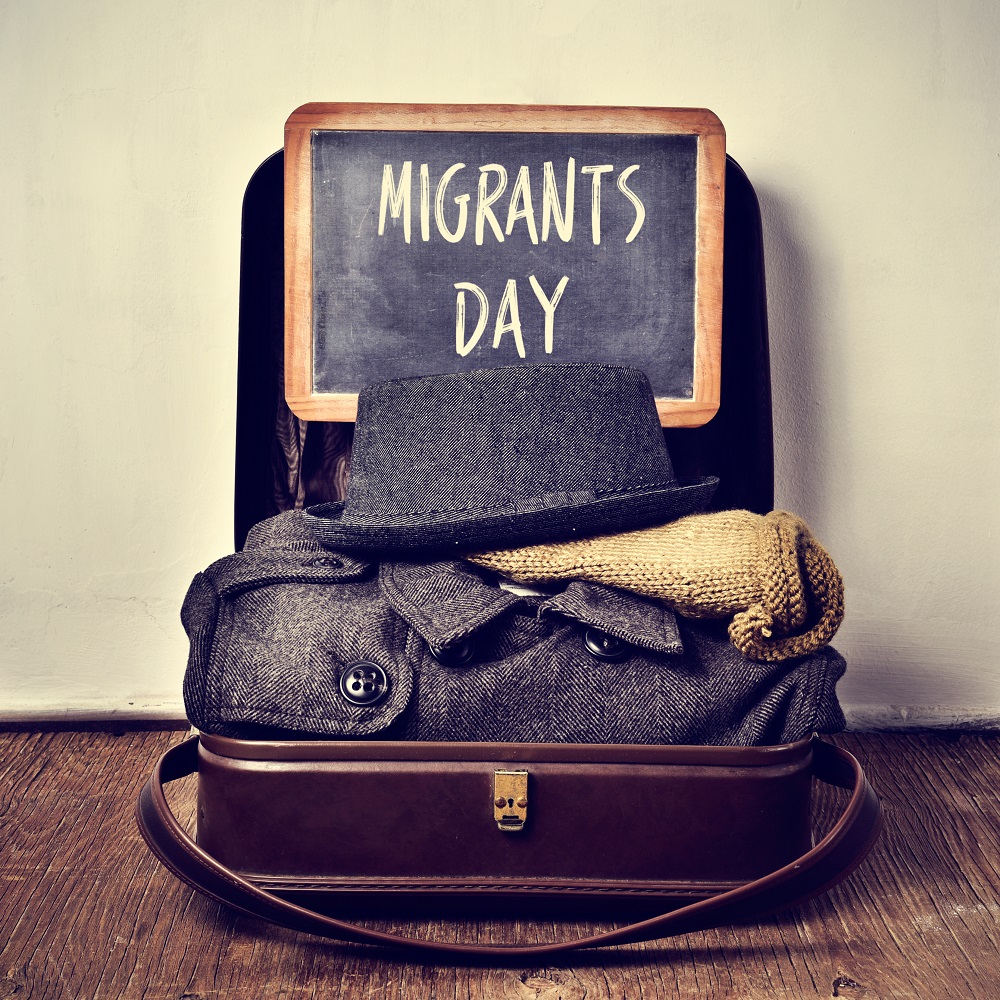 old suitcase with some clothing and chalkboard with text migrants day